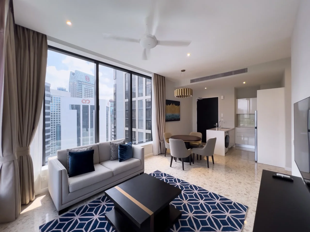 Living features: floor-to-ceiling windows, grey fabric sofa, black coffee table, area rug with geometric pattern, 3-blade ceiling fan, wall-mounted TV, decorative wall art, curtains, light-colored walls, marble flooring; style: modern; view: city. Kitchen features: white cabinetry, stainless-steel fridge, built-in oven, countertop with bar seating, open layout; style: modern.